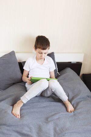 Distance learning online education. Boy sitting with a tablet on the bed in the room. Child with a gadget. Quarantine and self-isolation.の写真素材