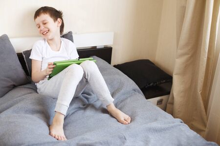 Distance learning online education. Boy sitting with a tablet on the bed in the room. Child with a gadget. Quarantine and self-isolation.の写真素材