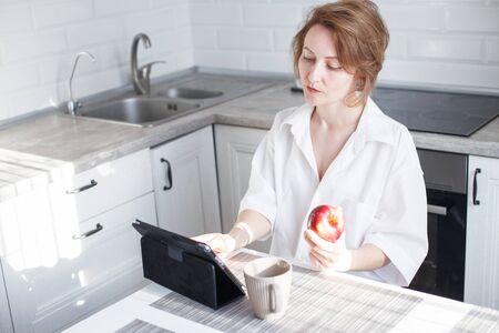 Happy beautiful woman with cup of coffee or tea using laptop in quarantine lockdown in the kitchen at home in the white shirt. Social distancing Self Isolationの写真素材