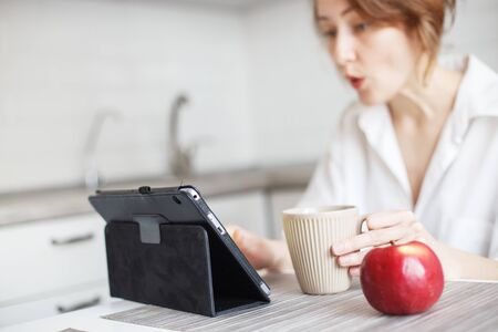 Happy surprised woman with cup of coffee or tea using laptop in quarantine lockdown in the kitchen...