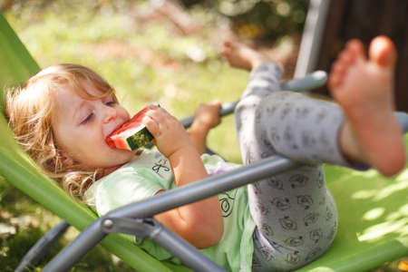 cute little girl eating watermelon on a deck chair in the garden in summertimeの写真素材