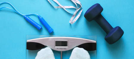 Woman measures her weight by standing her feet in socks on scale on a blue background with skipping rope and dumbbells. Flat lay composition. Healthy active lifestyleの写真素材