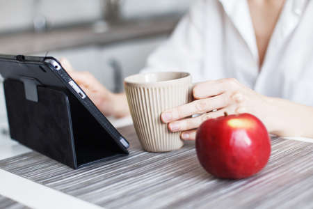 l woman with cup of coffee or tea using laptop in quarantine lockdown in the kitchen at home in the white shirt. girl with a tabletの写真素材