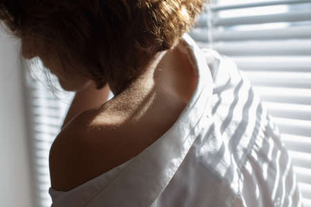 Sensual close up portrait of a beautiful young lady relaxing at the window. female shoulder close up. hard shadowsの写真素材