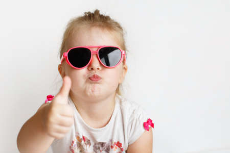 Child in pink sunglasses making funny face. positive girl thumbs up on white backgroundの写真素材