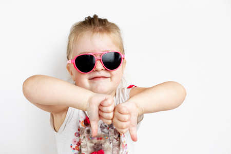 Child in pink sunglasses making funny face. positive girl thumbs down on white backgroundの写真素材