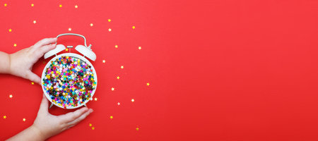 child's hands hold a white alarm clock with scattered sequins in the form of stars on a red background. Party, christmas, new year, celebration concept. copy space. Flat lay. Top view. bannerの写真素材