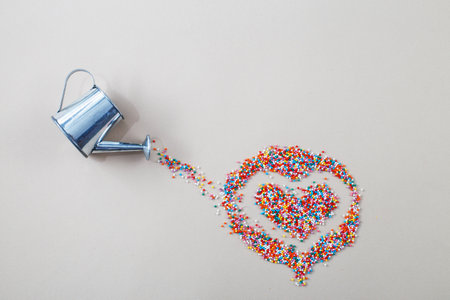 silver watering can pouring sugar baking colorful sprinkles in shape of heart on a light beige background.の写真素材