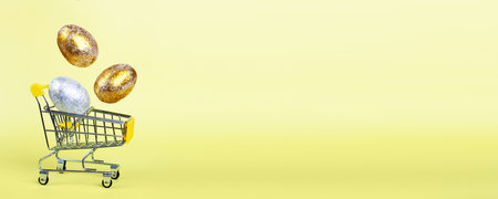 Easter golden and silver eggs levitation to the shopping cart on a yellow background. Easter concept. Copy space. bannerの写真素材