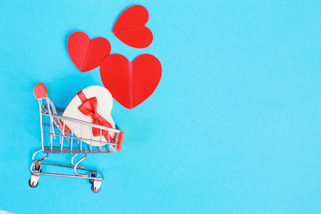 Shopping cart with a red heart and gift box on blue background. Shopper accessory, consumerism and commerce symbolの写真素材