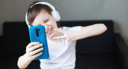 leisure, children, technology and people concept - smiling boy with smartphone and headphones having video call at home. child with mobile. bannerの写真素材