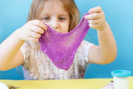Blonde girl hold and stretching purple glitter slime isolated on a blue background. child playing with a slime toy. Making slime. Copy space. DIY concept.の写真素材