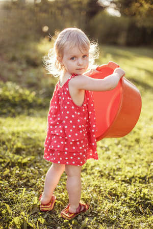 A cute baby girl in the red dress hold plastic basin outdoor.の写真素材