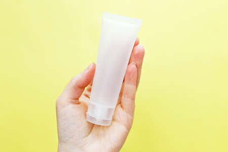 Female hand holding cream tube cosmetic products on a yellow background.
