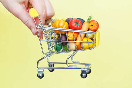 Metal shopping cart with fruits and vegetables on the persons hand on yellow background. Food delivery internet shopping conceptの写真素材