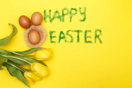 inscription happy easter from grass with bunny eggs, flowers tulips and nest on a yellow background. copy space for textの写真素材