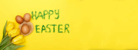 inscription happy easter from grass with bunny eggs, flowers tulips and nest on a yellow background. copy space for text. bannerの写真素材