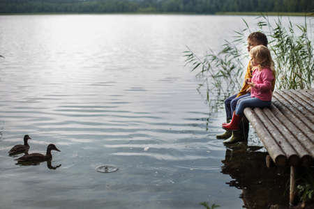 Brother and sister sitting on the river pier and hug and look at ducks in the water friendship concept. Happy cute kids feed ducksの写真素材