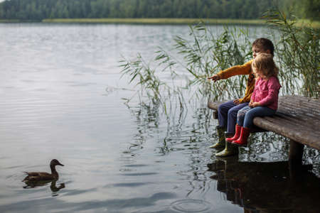 Brother and sister sitting on the river pier and hug and look at ducks in the water friendship concept. Happy cute kids feed ducksの写真素材