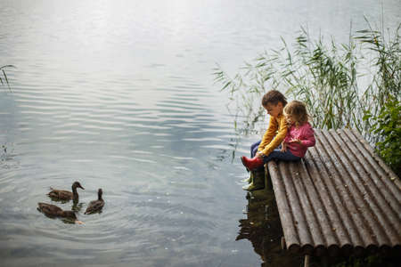 Brother and sister sitting on the river pier and hug and look at ducks in the water friendship concept. Happy cute kids feed ducksの写真素材