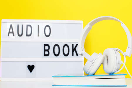 Audio book concept with modern white headphones and hardcover book on a yellow background. Listening to a book. E-learning. Copyspace. inscription on a lightboxの写真素材