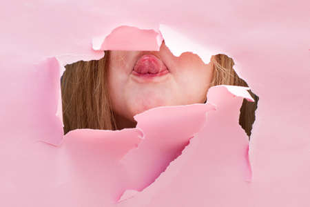 portrait of a funny kid girl showing tongue in a torn paper hole on a pink backgroundの写真素材