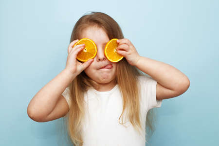little happy girl with halves of oranges covering eyes on a blue background. child having fun and showing tongue.の写真素材