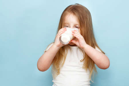 little beautiful girl drinking milk in a glass on a blue backgroundの写真素材