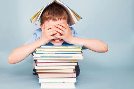 Upset schoolboy sitting with pile of school books and covers his face with hands isolated on a blue backgroundの写真素材
