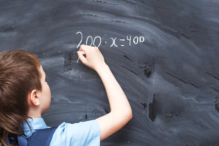 Boy standing back in front of a school blackboard and writing. Schoolboy solves math example at the...