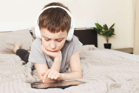 child boy lying on bed at home with pc tablet and reading online or learning or listening to music with wireless headphonesの写真素材