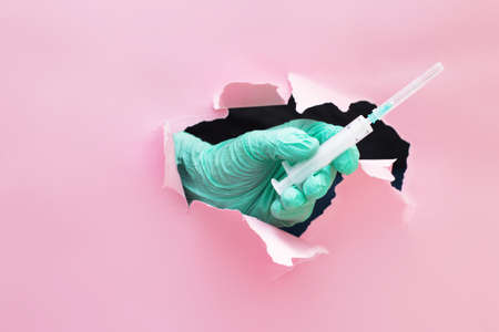 a doctor's hand holds out a syringe with a vaccine through a hole in torn paper on a pink backgroundの写真素材