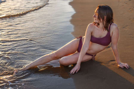 a pretty brunette girl in bikini sits in the water on the seaside and enjoying the sunsetの写真素材