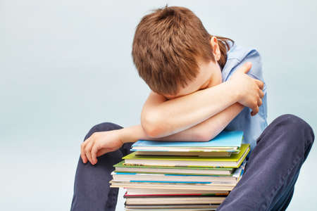 Upset schoolboy sitting with pile of school books and covers his face with hands. boy sleeping on a stack of textbooks isolated on a blue backgroundの写真素材