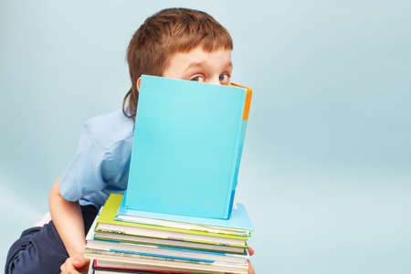 Schoolboy is sitting with a stack of books and reading and covers his face with a book isolated on a blue backgroundの写真素材