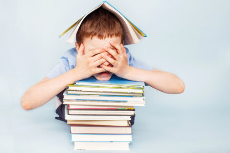 Upset schoolboy sitting with pile of school books and covers his face with hands isolated on a blue backgroundの写真素材
