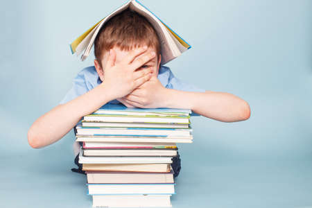 Upset schoolboy sitting with pile of school books and covers his face with hands isolated on a blue backgroundの写真素材