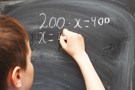 Boy standing back in front of a school blackboard and writing. Schoolboy solves math example at the chalkkboardの写真素材
