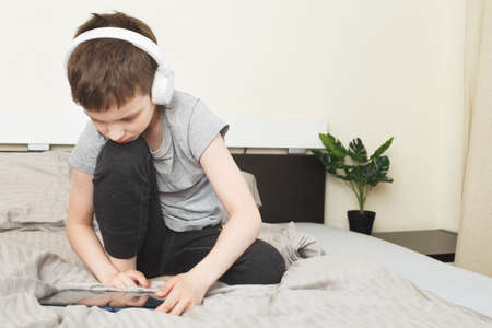child boy sitting on bed at home with pc tablet and reading online or learning or listening to music with wireless headphonesの写真素材