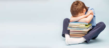 Upset schoolboy sitting with pile of school books and covers his face with hands. boy sleeping on a stack of textbooks isolated on a blue background. bannerの写真素材