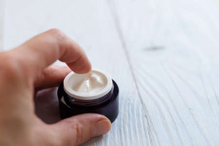 Skin and body care. Hands of a girl with a jar of face cream close up applied to the finger of woman's hand on light blue wooden backgroundの写真素材
