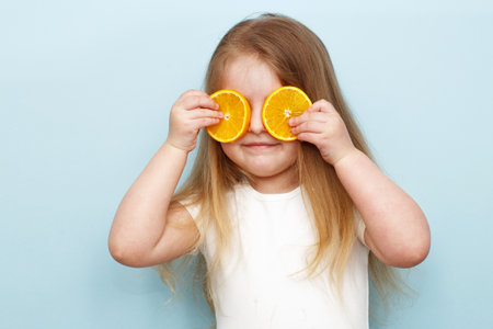 little happy girl with halves of oranges covering eyes on a blue backgroundの写真素材