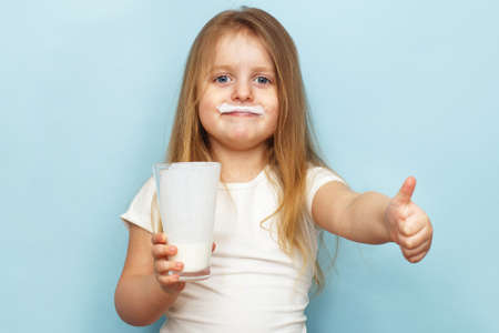 little beautiful girl holding milk in a glass on a blue background. child drinks yogurtの写真素材