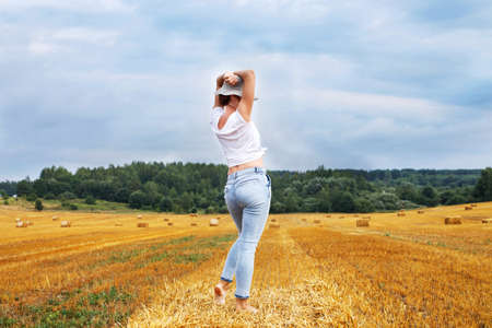 barefoot girl in straw hat stands on a haystack on a bale in the agricultural field after harvesting.の写真素材