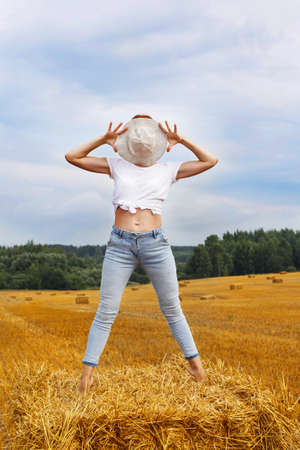 barefoot girl in straw hat stands on a haystack on a bale in the agricultural field after harvesting.の写真素材