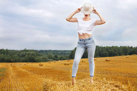 barefoot girl in straw hat stands on a haystack on a bale in the agricultural field after harvesting.の写真素材