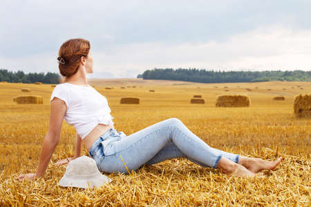 barefoot girl with straw hat sitting on a haystack on a bale in the agricultural field after harvesting.の写真素材