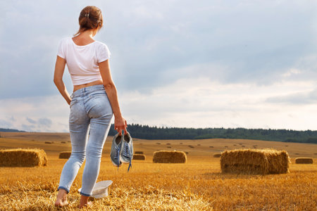 barefoot girlwith sneakers in hand stands on a haystack on a bale in the agricultural field after harvesting.の写真素材