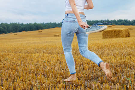 barefoot girl with sneakers in hand walking in the agricultural field with haystack and bales after harvesting.の写真素材