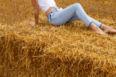 barefoot girl sitting on a haystack on a bale in the agricultural field after harvesting.の写真素材